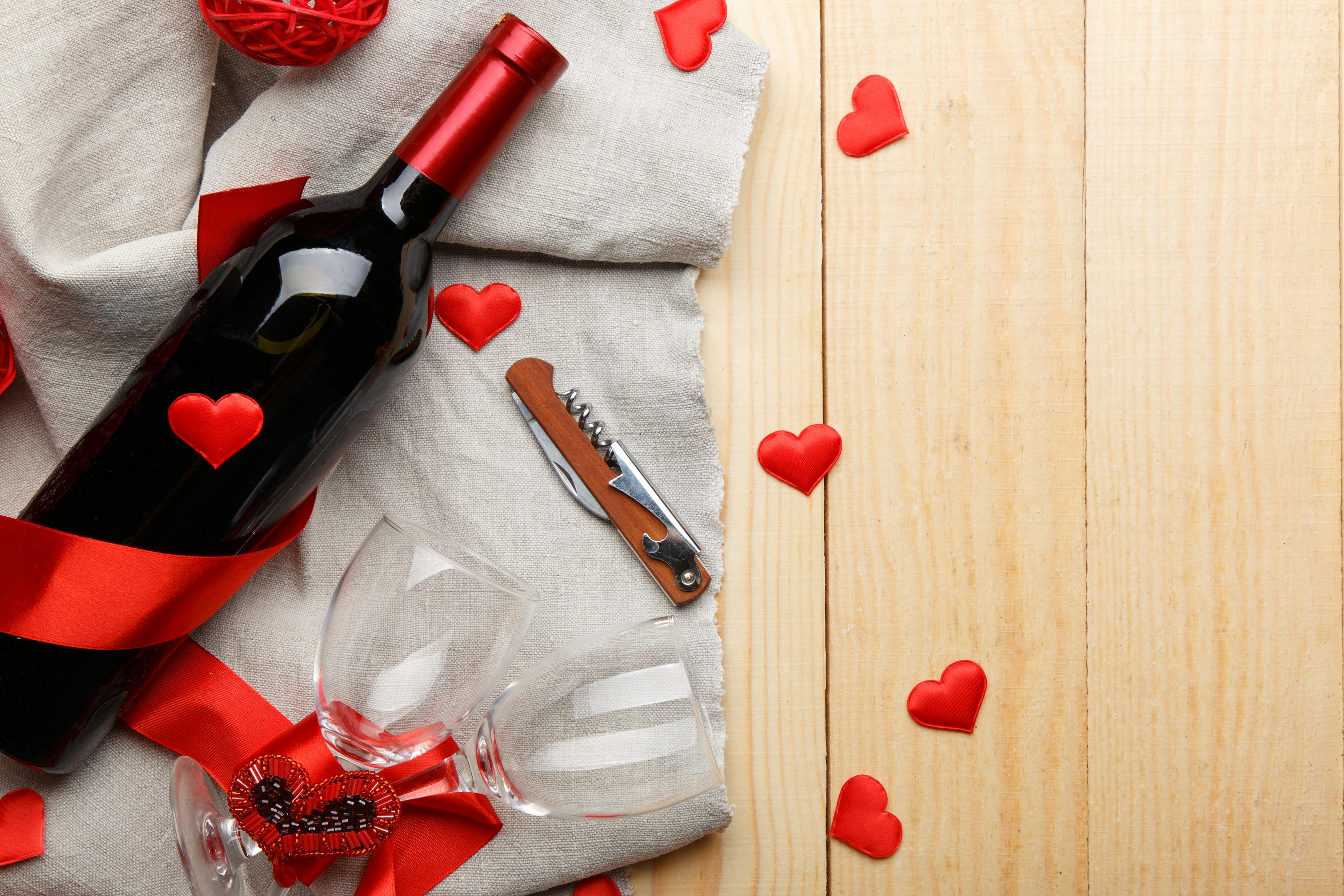 A romantic flat lay of wine bottle, glasses, and red hearts on a wooden surface with burlap.