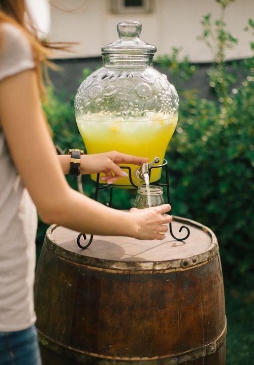 Free Woman pouring fresh lemonade from a dispenser on a wooden barrel in a sunny Turkish garden. Stock Photo