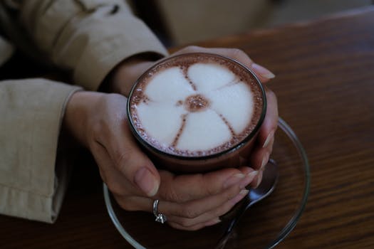 Hands holding a cup of latte with delicate milk foam art on a wooden table.