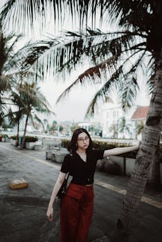 Stylish young woman posing under palm trees in a vibrant urban setting.