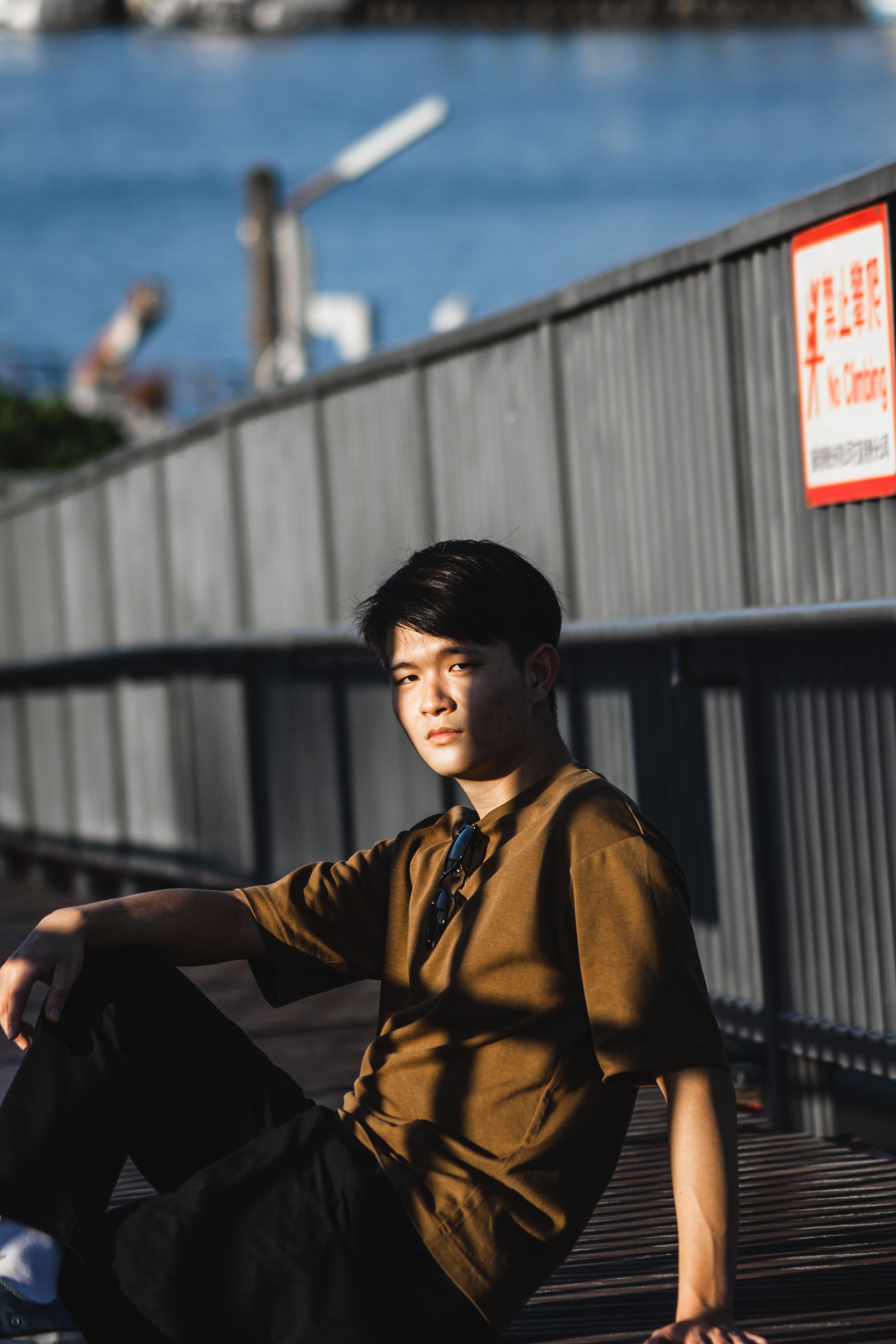 Asian man in casual attire sitting on a boardwalk with a waterfront view.