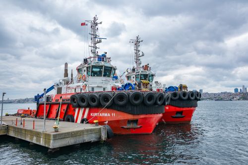 Free Two red tugboats docked under a cloudy sky at an urban harbor, ready for action. Stock Photo
