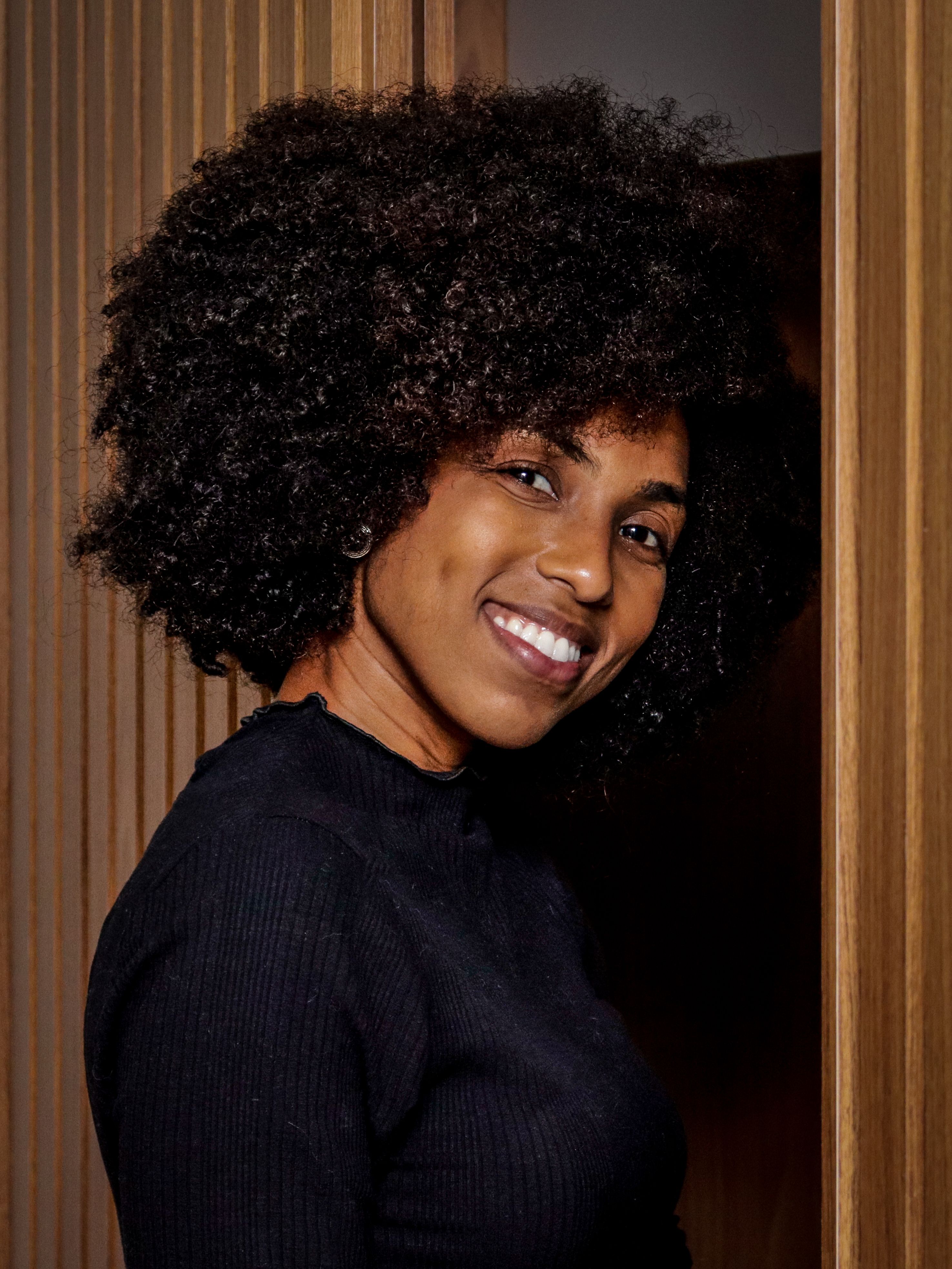 Smiling woman with curly afro hairstyle in a wooden interior setting in Niterói, Brazil.