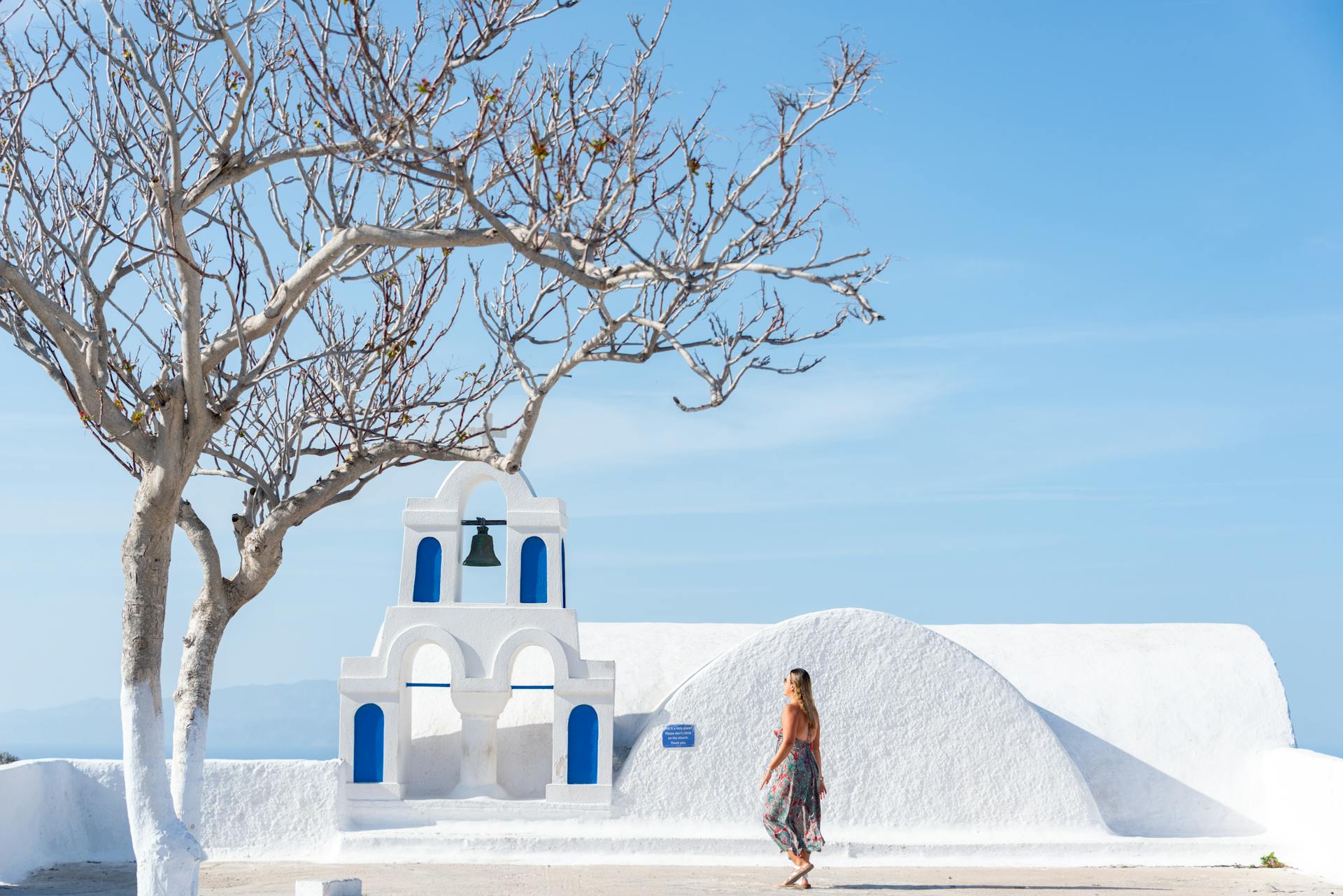 Woman strolling near Santorini's iconic bell tower under clear blue skies.