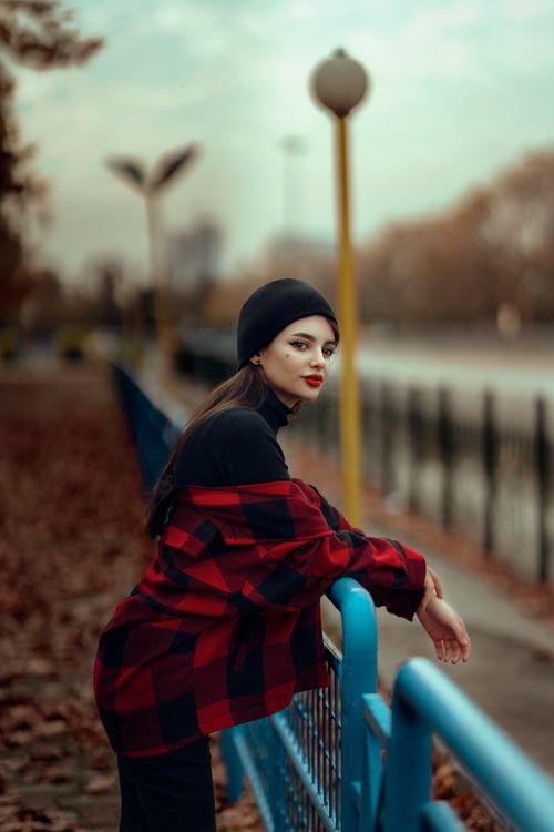Free Woman leans casually on a blue railing in autumn, wearing a stylish red plaid jacket and black beanie. Stock Photo