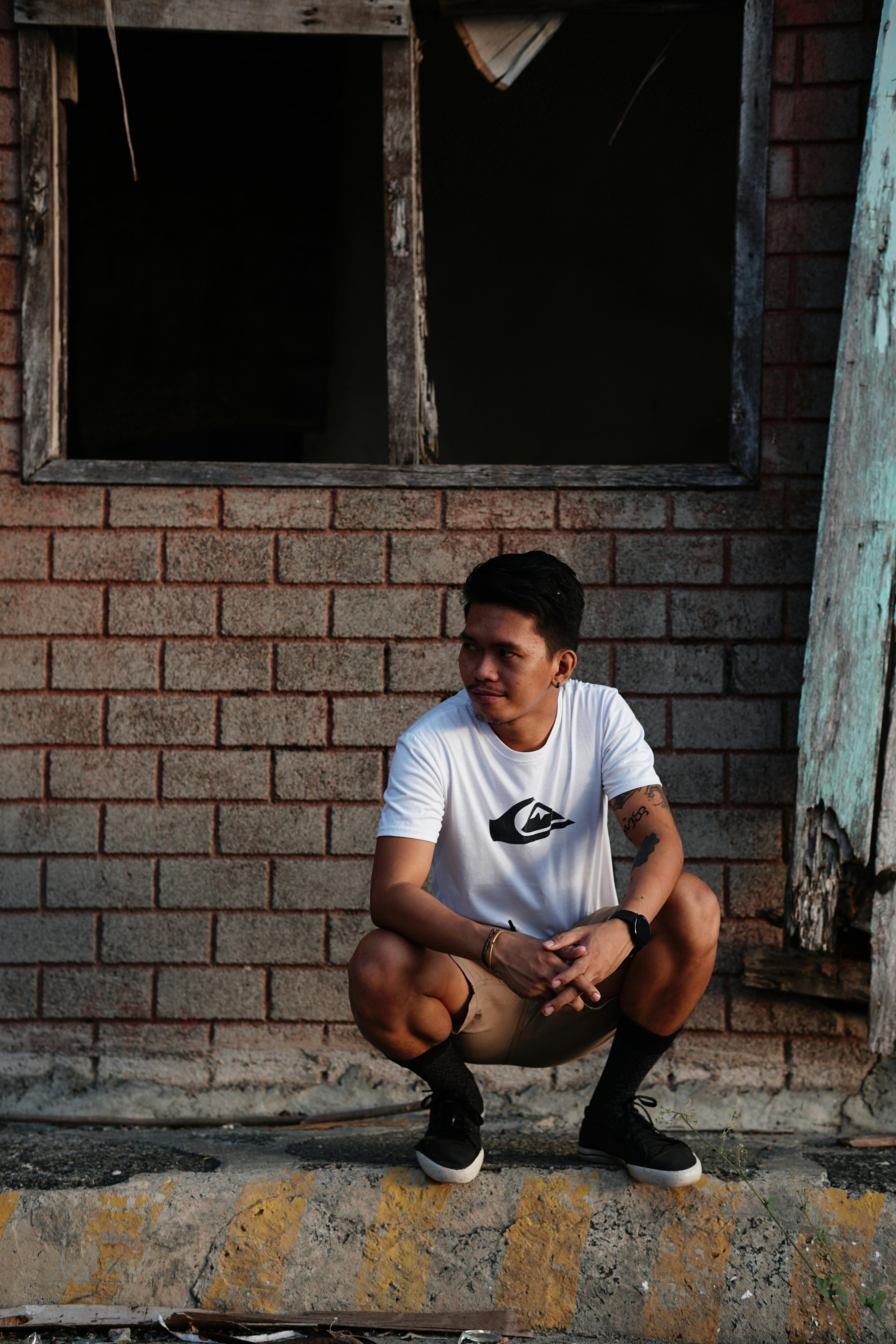 Casual photo of a man in shorts crouching against a decaying brick wall outdoors.