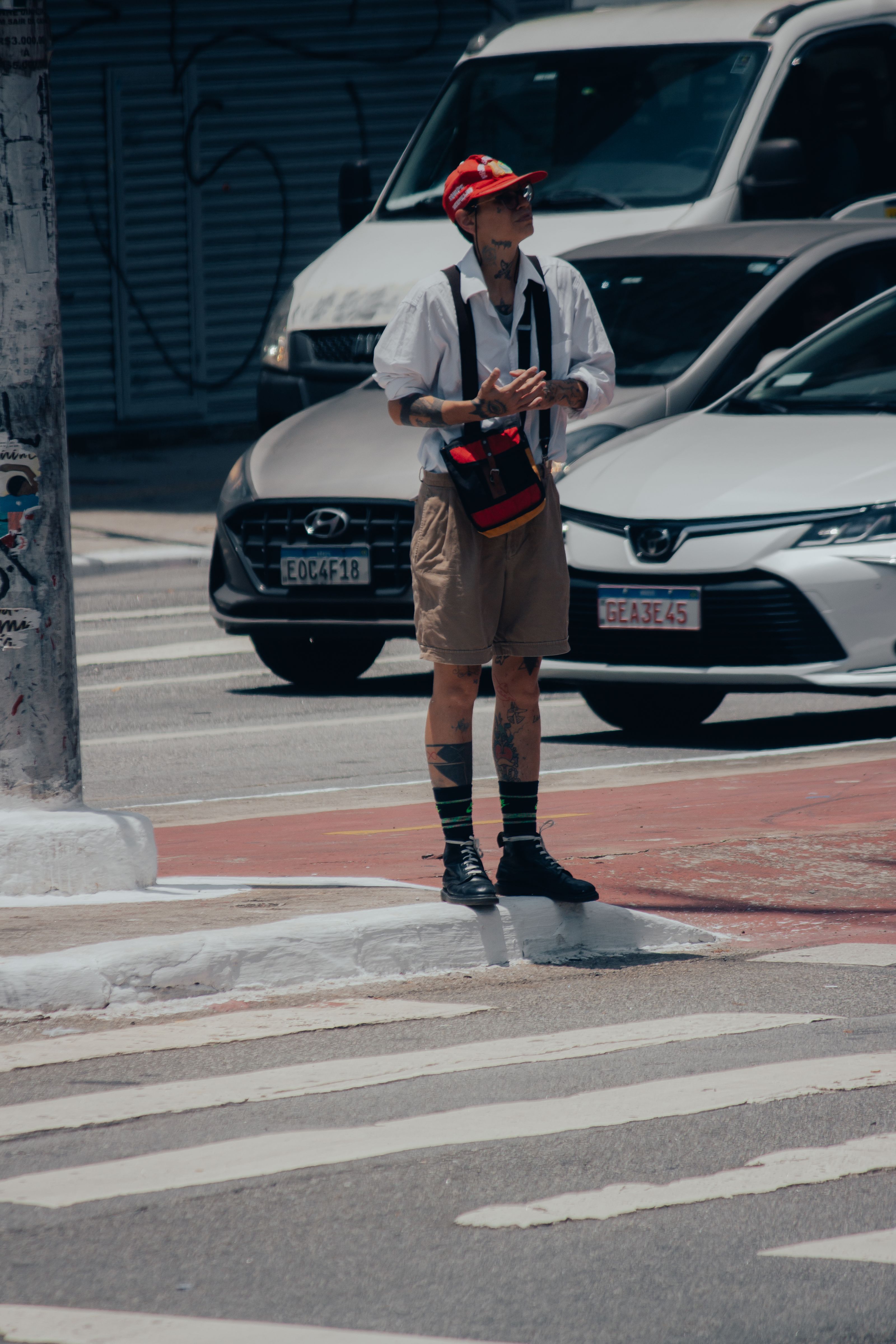 Um homem com tatuagens e um boné espera na faixa de pedestres em um ambiente urbano em um dia ensolarado.