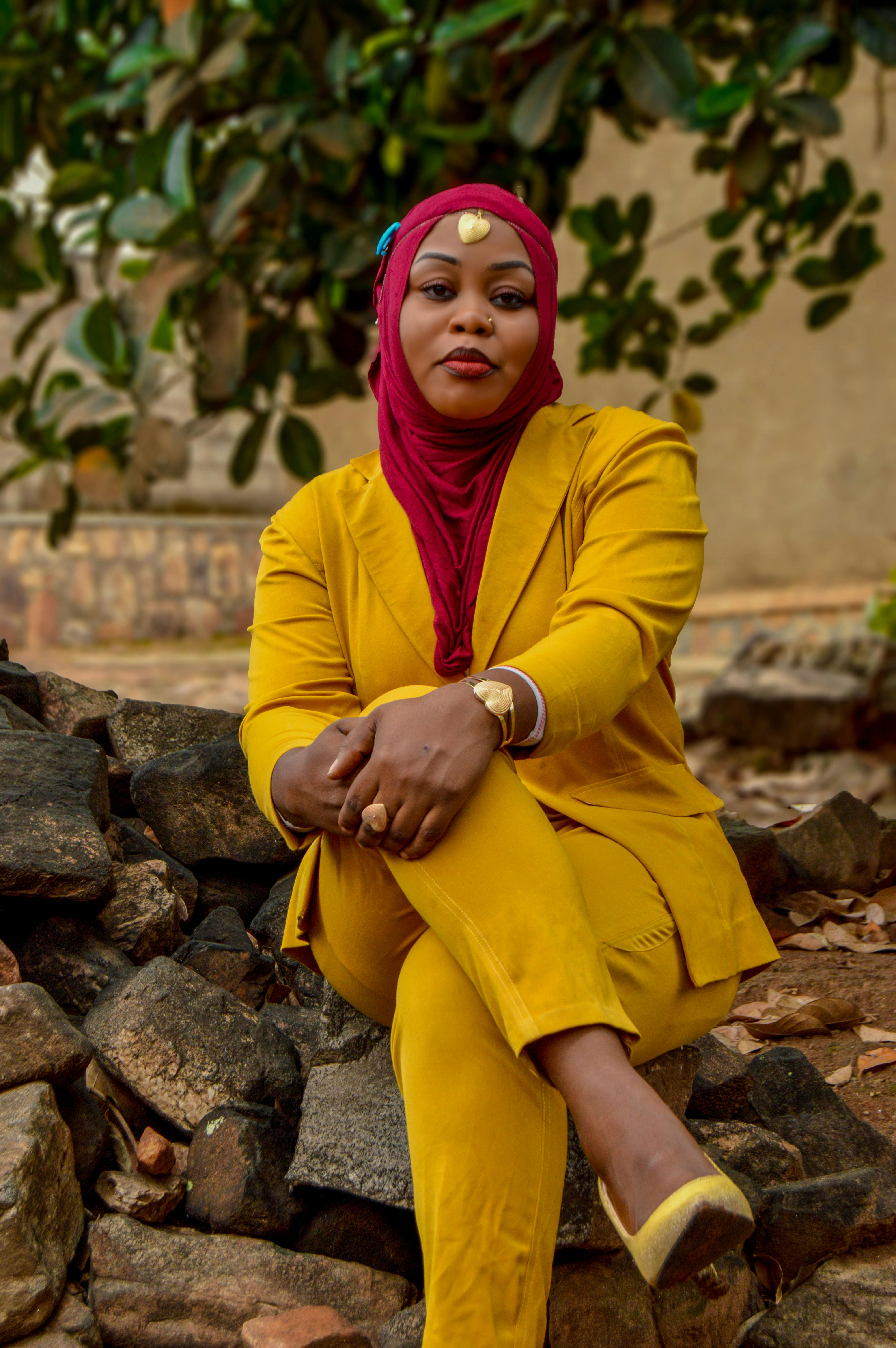 Stylish woman in hijab and yellow suit sitting on rocks outdoors.