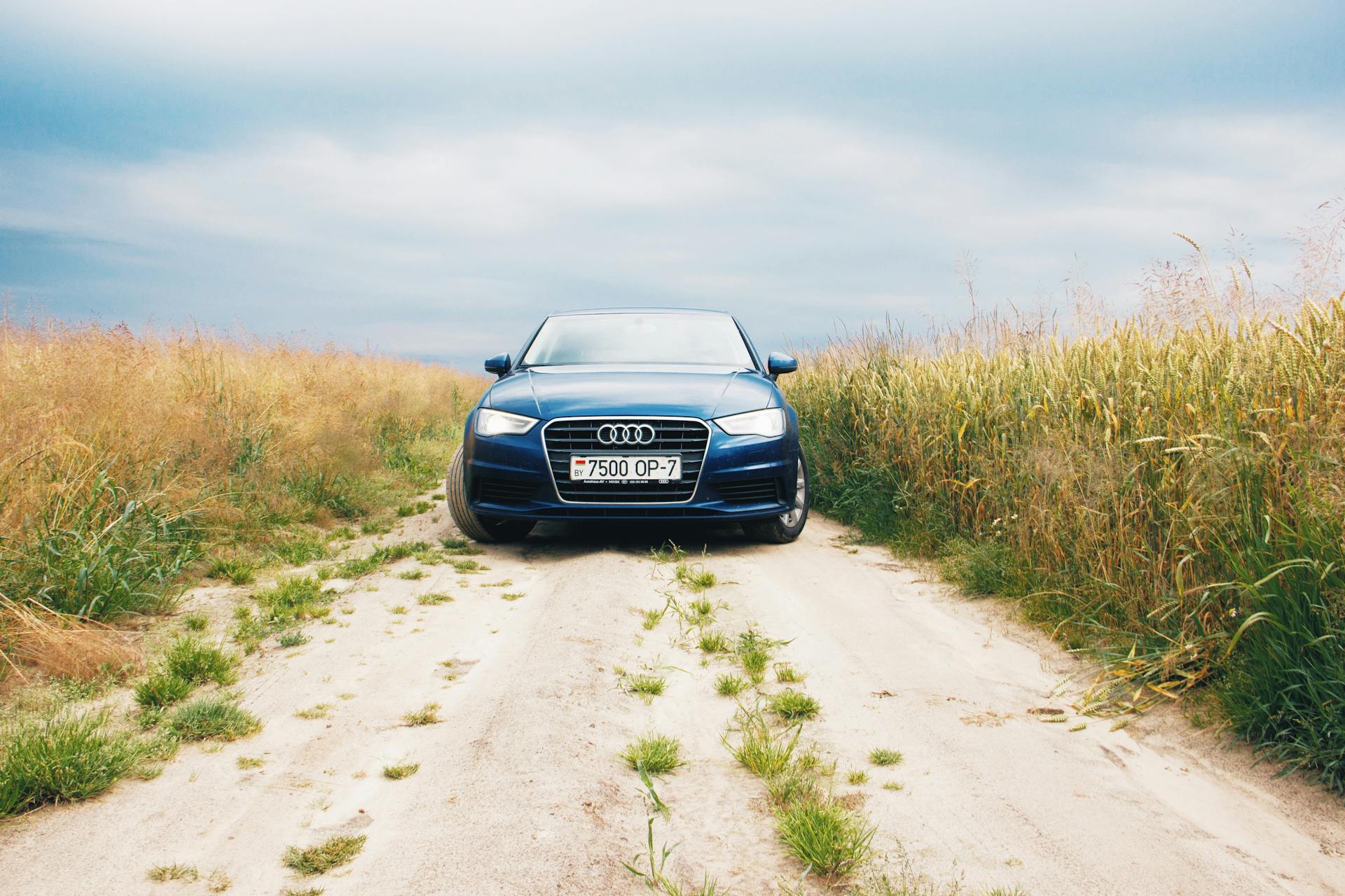 A luxury car parked on a rural sandy road surrounded by fields under a cloudy sky.