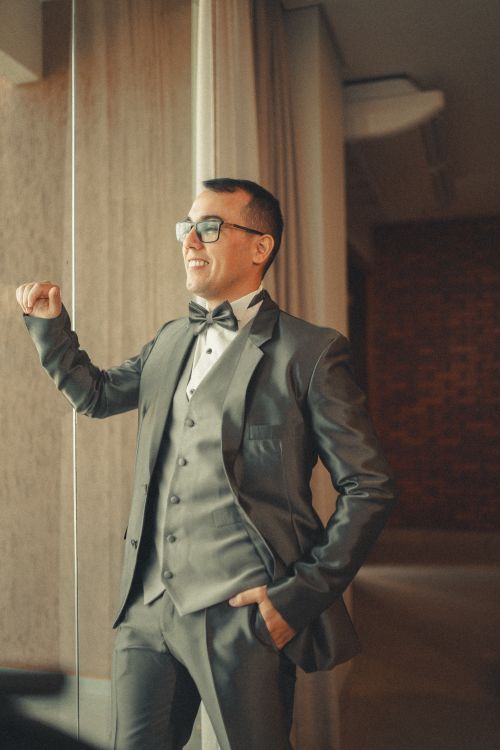 Free A groom stands smiling in a stylish gray suit with a bowtie indoors, capturing the essence of wedding photography. Stock Photo