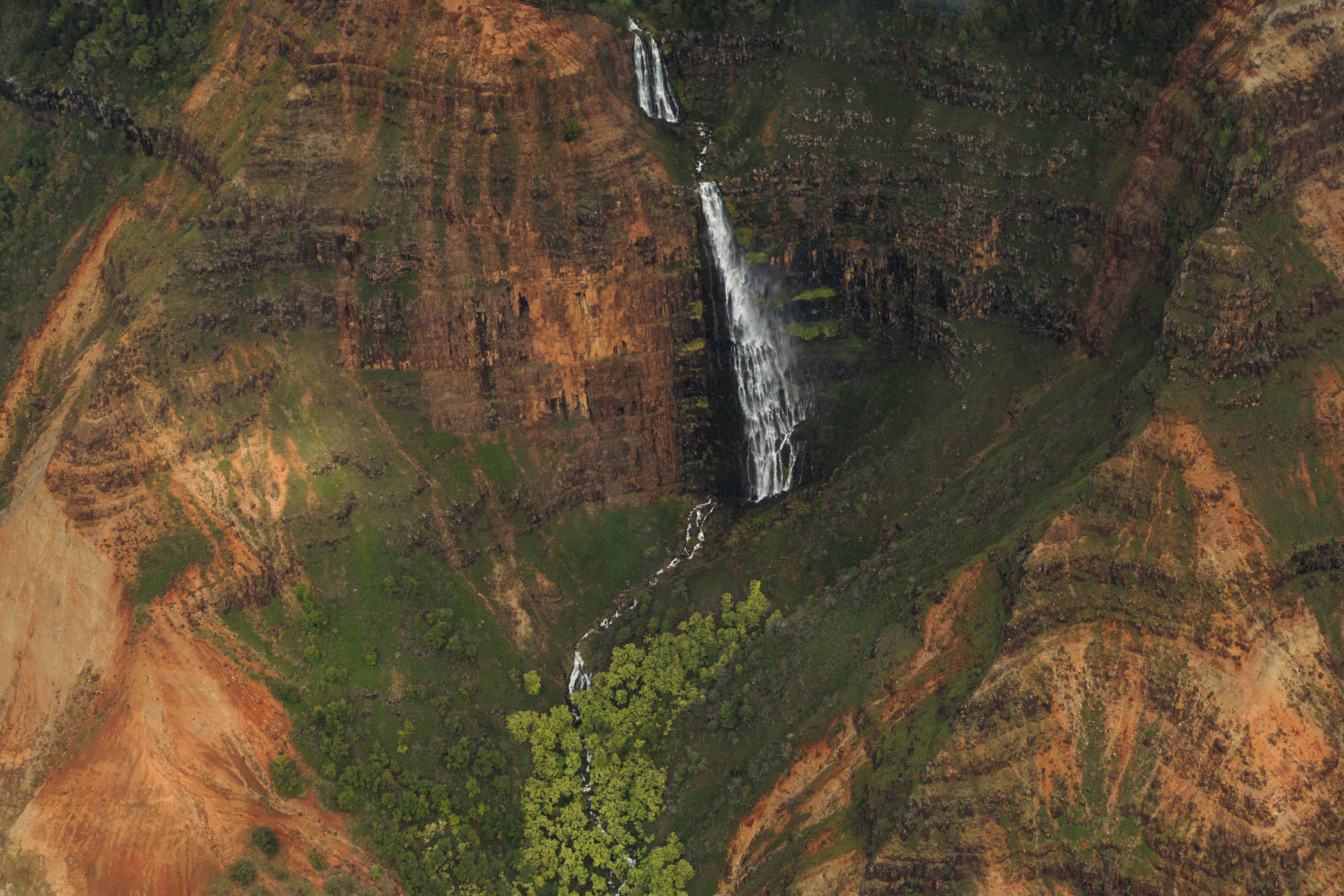 KAUAI LUXURY 60-Minute Doors-Off Helicopter ADVENTURE - Stunning aerial shot of a waterfall cascading down Waimea Canyon's vibrant rocky cliffs in Hawaii.