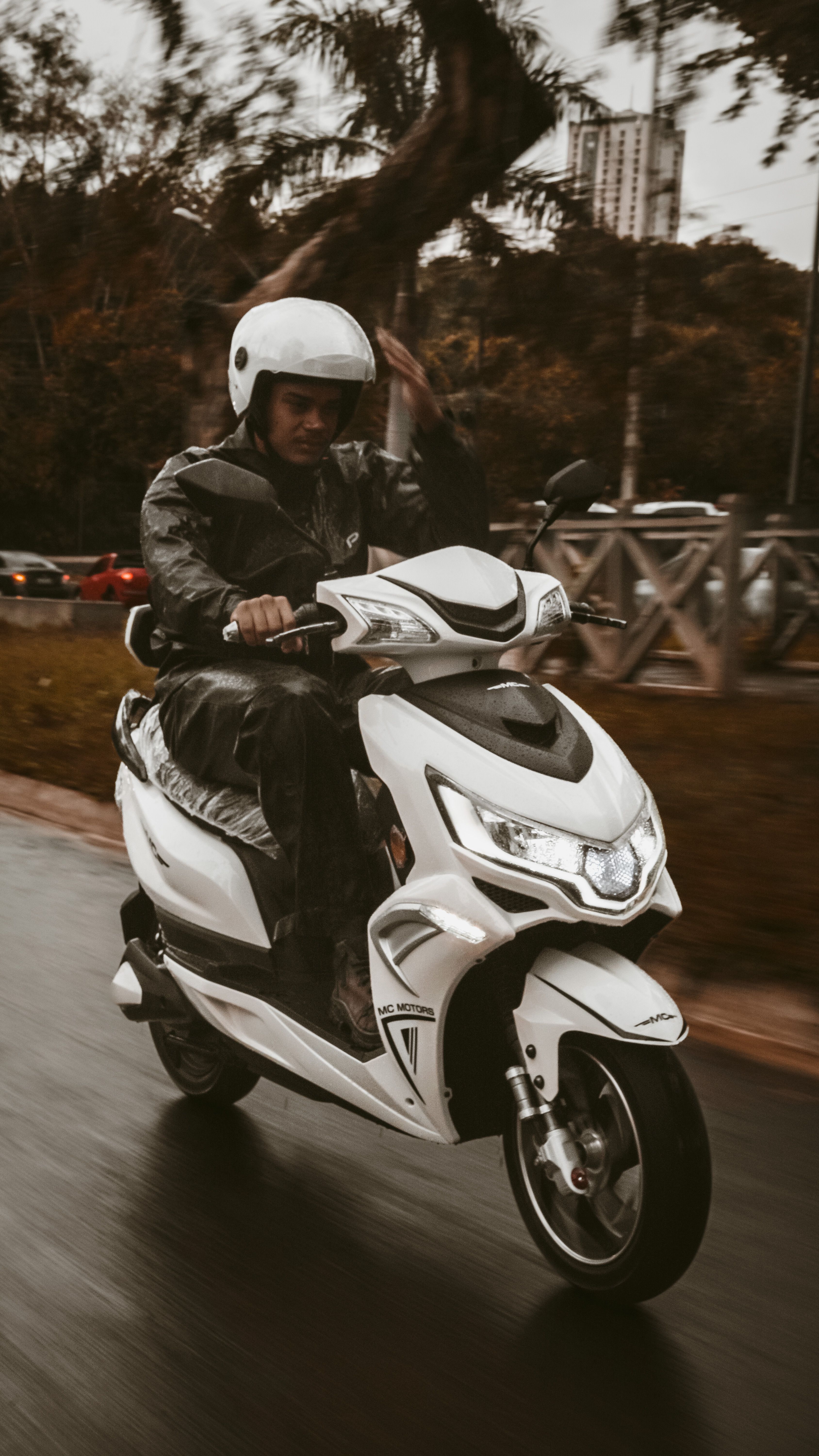 A man in a helmet rides a white scooter on a wet city road, capturing a dynamic urban scene.