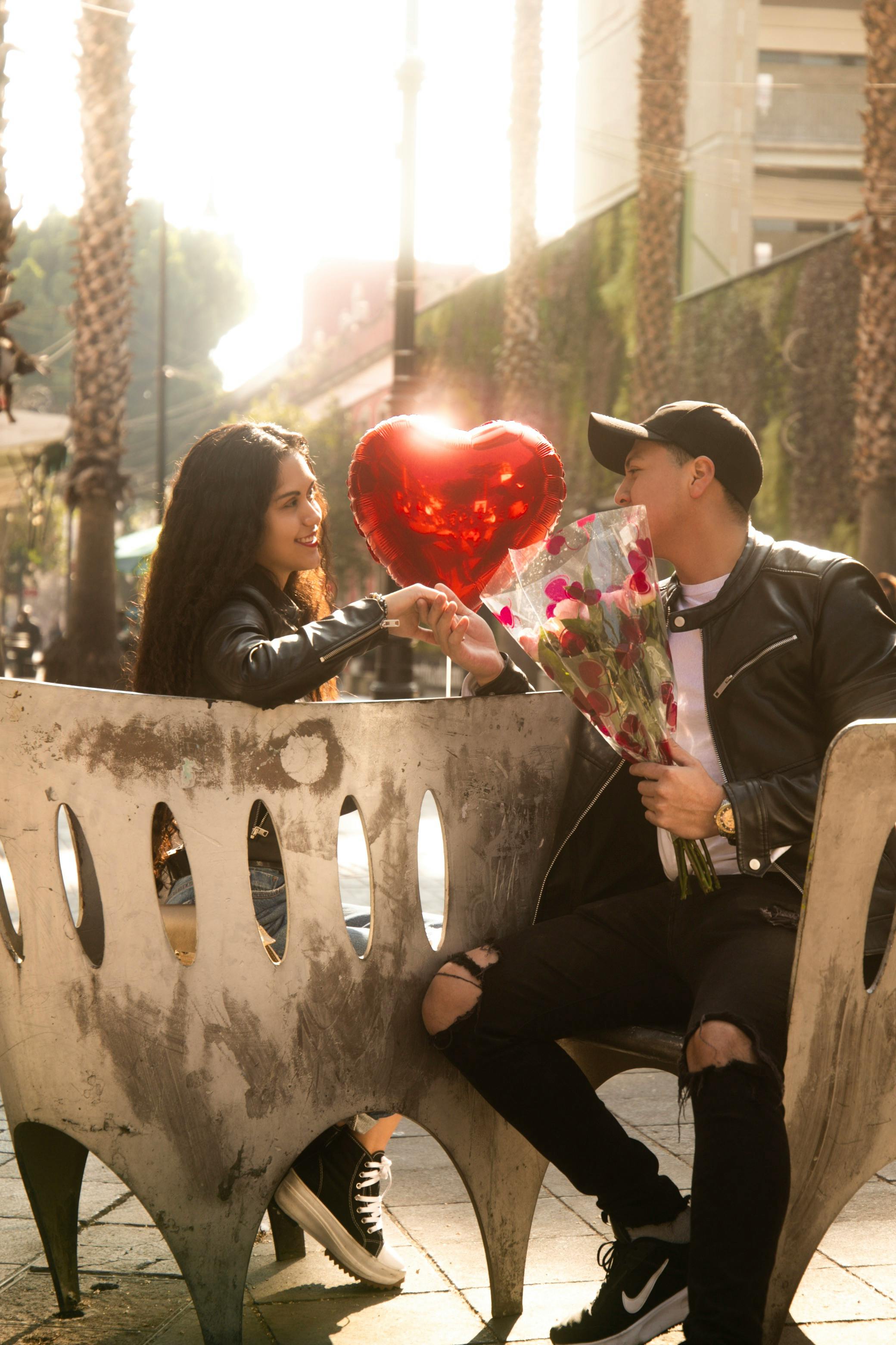 A couple sitting on a bench exchanging flowers and a heart balloon, enjoying a sunny day.