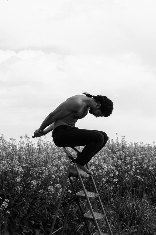 Free Artistic black and white photograph of a man sitting topless on a ladder in a meadow. Stock Photo