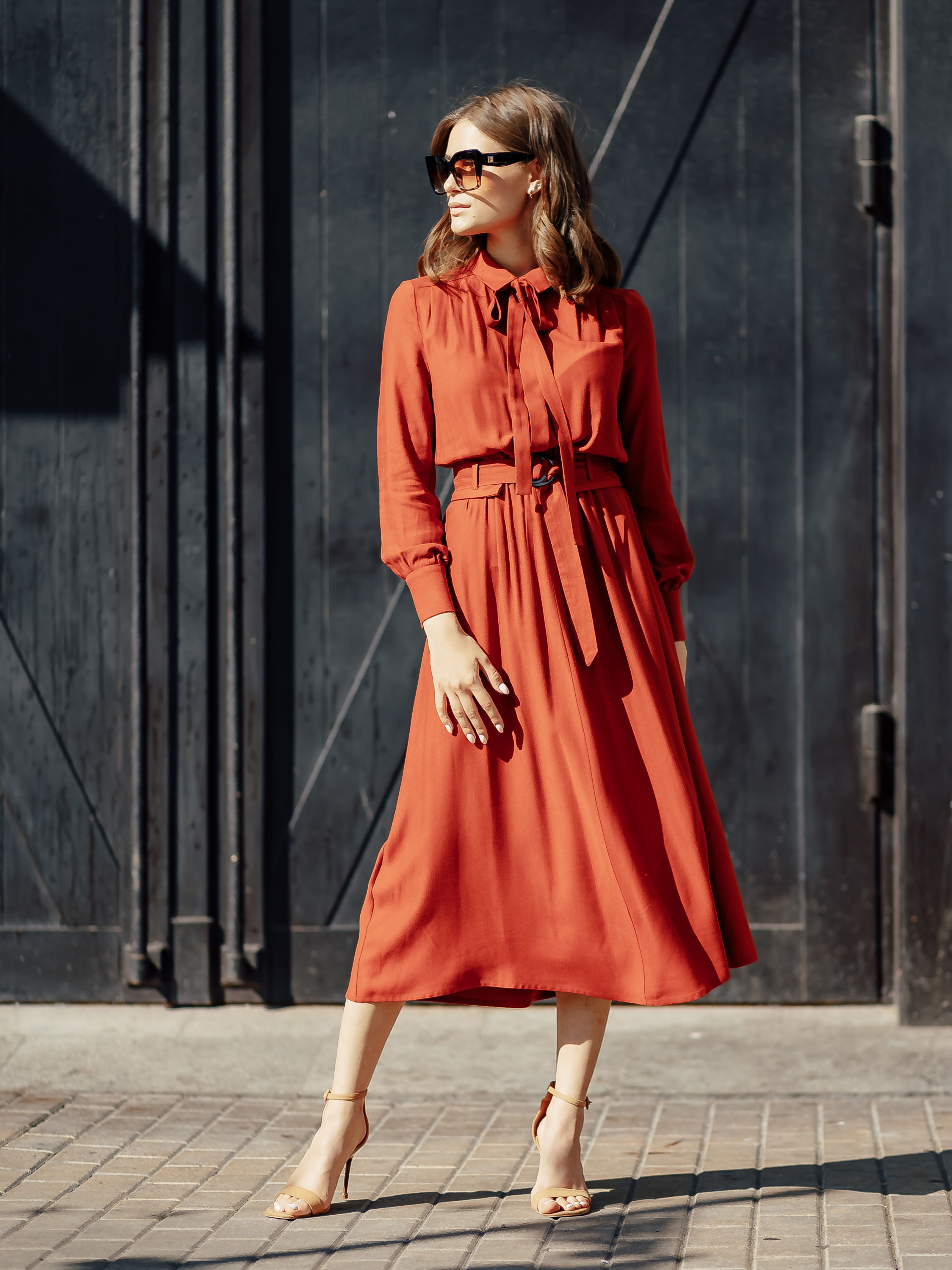 Mulher elegante em um vestido vermelho posando contra uma parede preta em um dia ensolarado.
