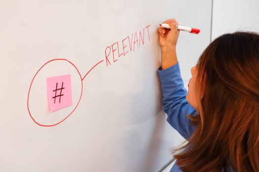 A woman writing the word "relevant" next to a sticky note with a hashtag on it