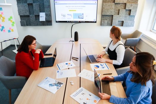 Three women collaborating in a modern office with laptops and presentation materials.