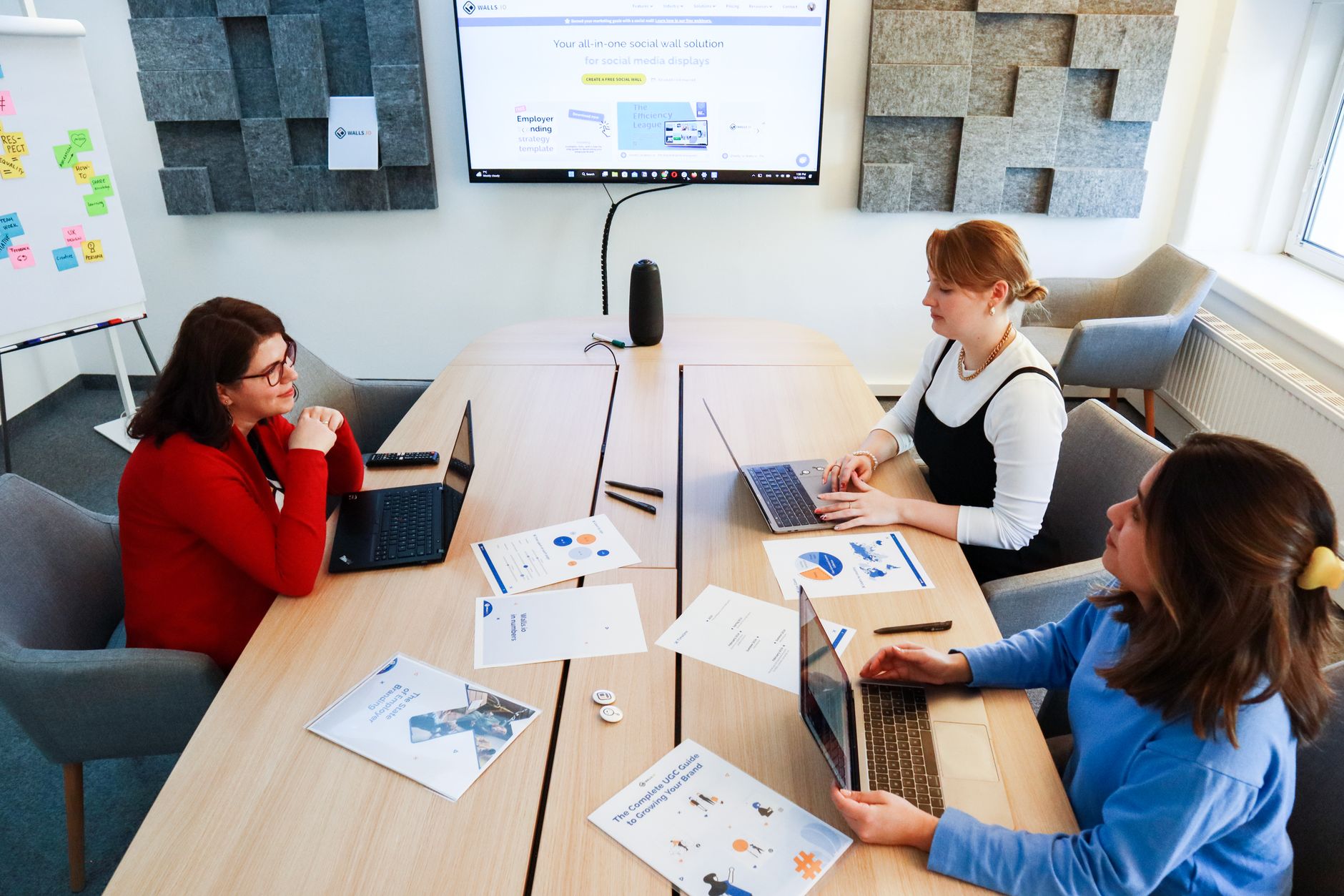 Three women discuss at a conference table in a modern office setting, using laptops and documents.