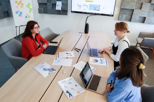 Three women in an office setting discussing business documents on laptops and paper.
