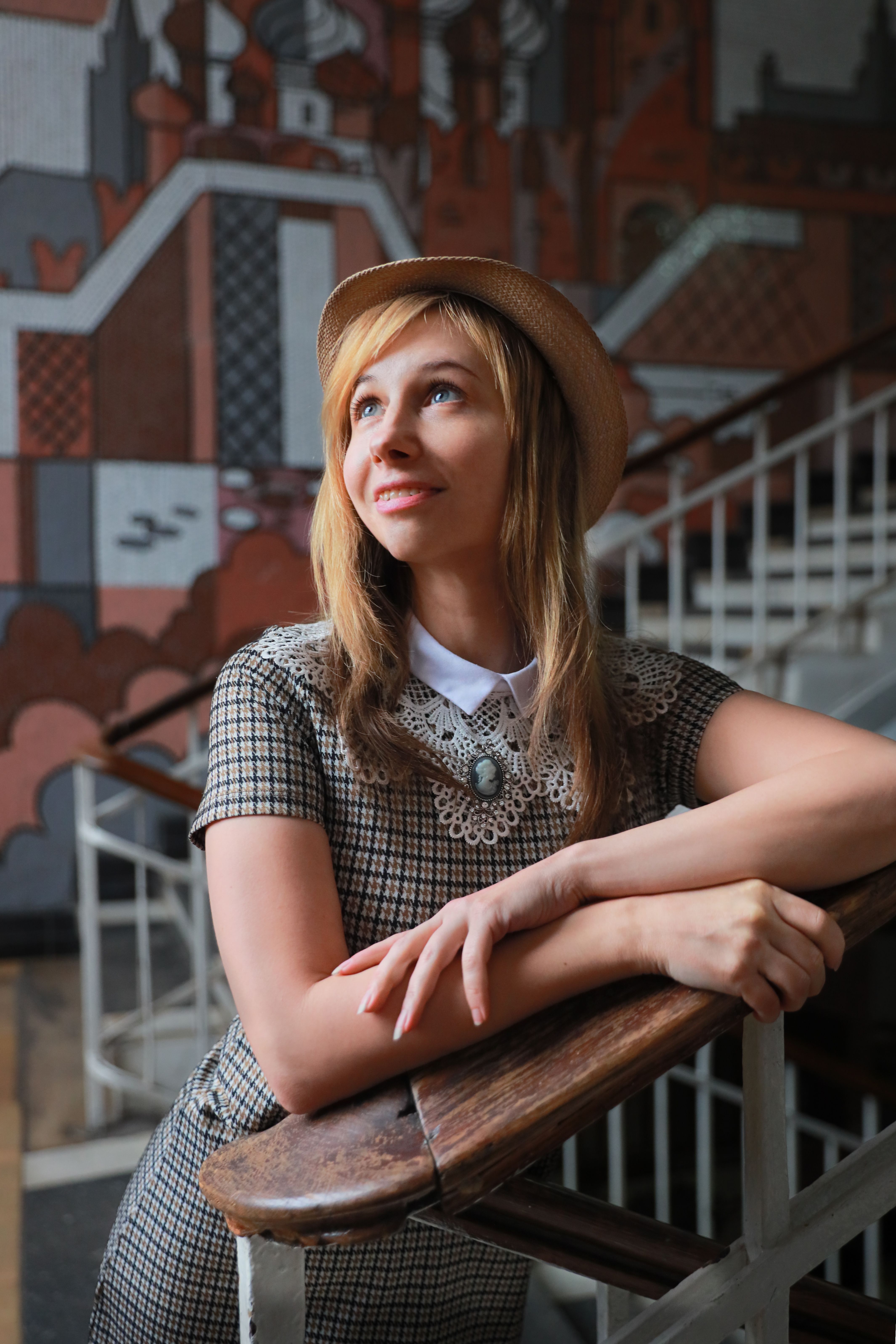 Portrait of a stylish young woman in a hat and dress posing on an ornate staircase indoors.