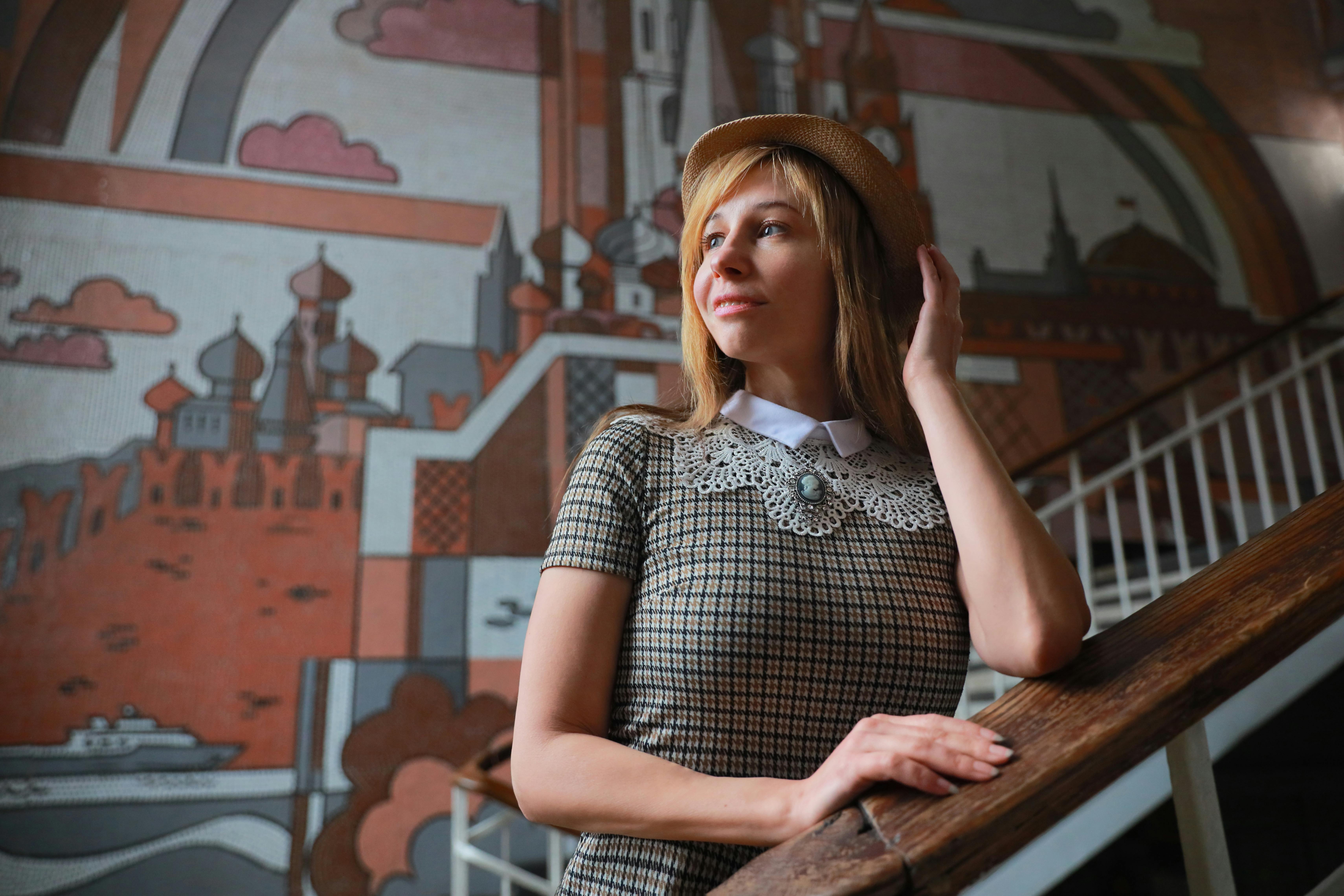 A stylish woman in a hat poses gracefully by a vibrant mural indoors.