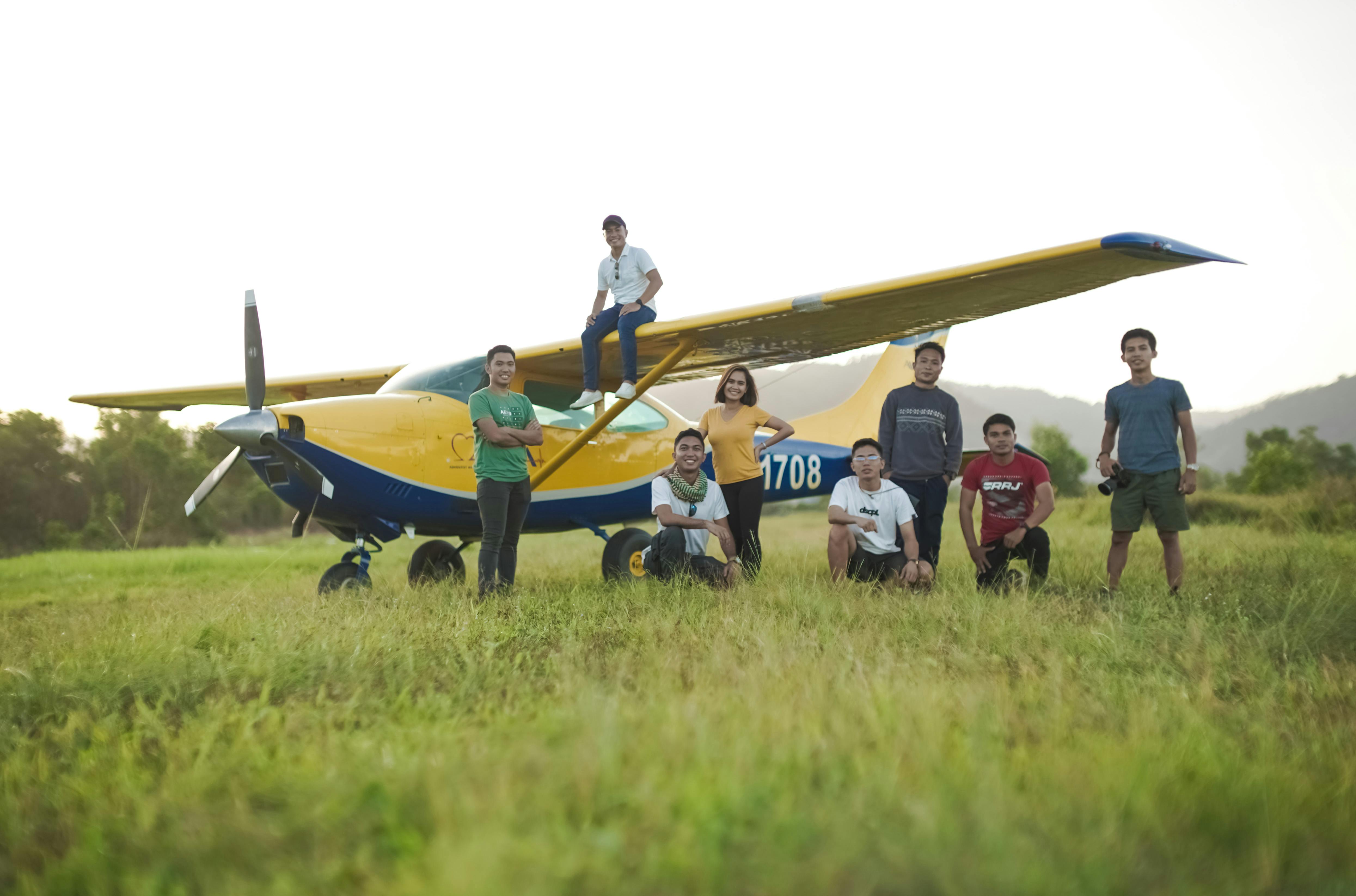 A group of young adults with a yellow Cessna airplane in a sunny meadow.