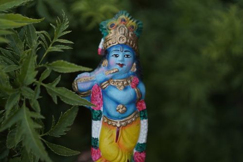 Free Vibrant handmade Krishna figurine surrounded by lush green leaves, showcasing intricate craftsmanship. Stock Photo