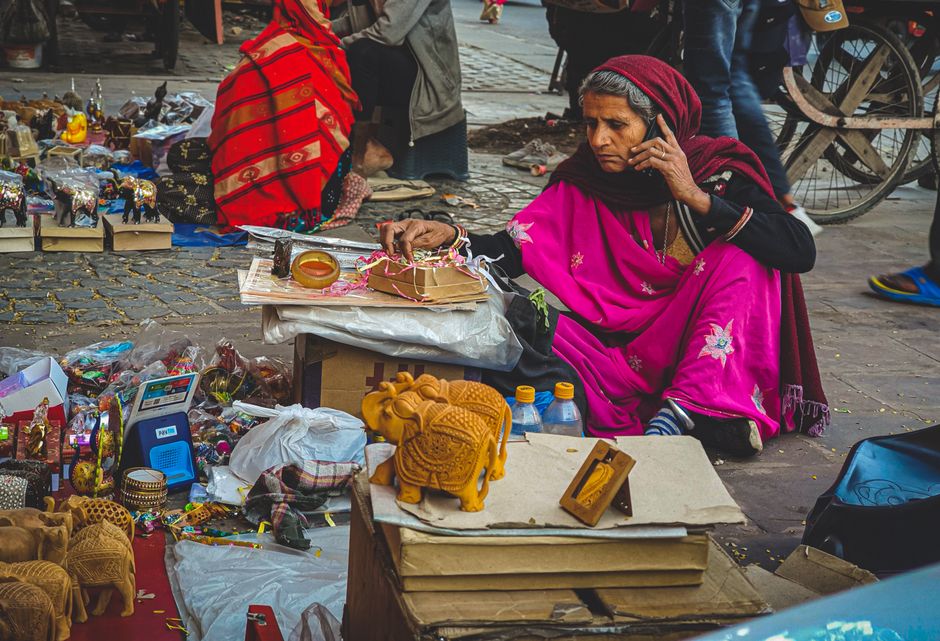 An elderly woman in traditional attire selling handicrafts at a Jaipur street market, talking on the phone.