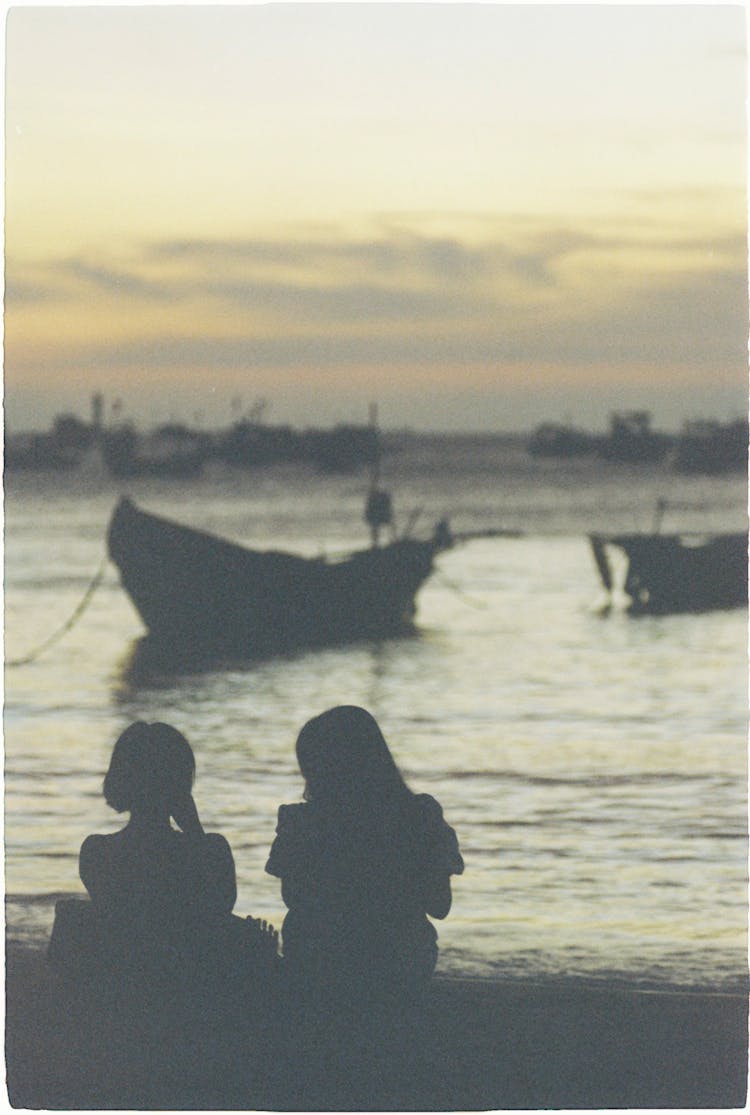 Silhouette Of Women Sitting On Shore At Sunset