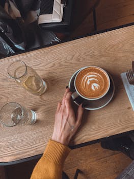 A top view of a hand holding a cup of latte with froth art on a wooden table, creating a warm cafe vibe.