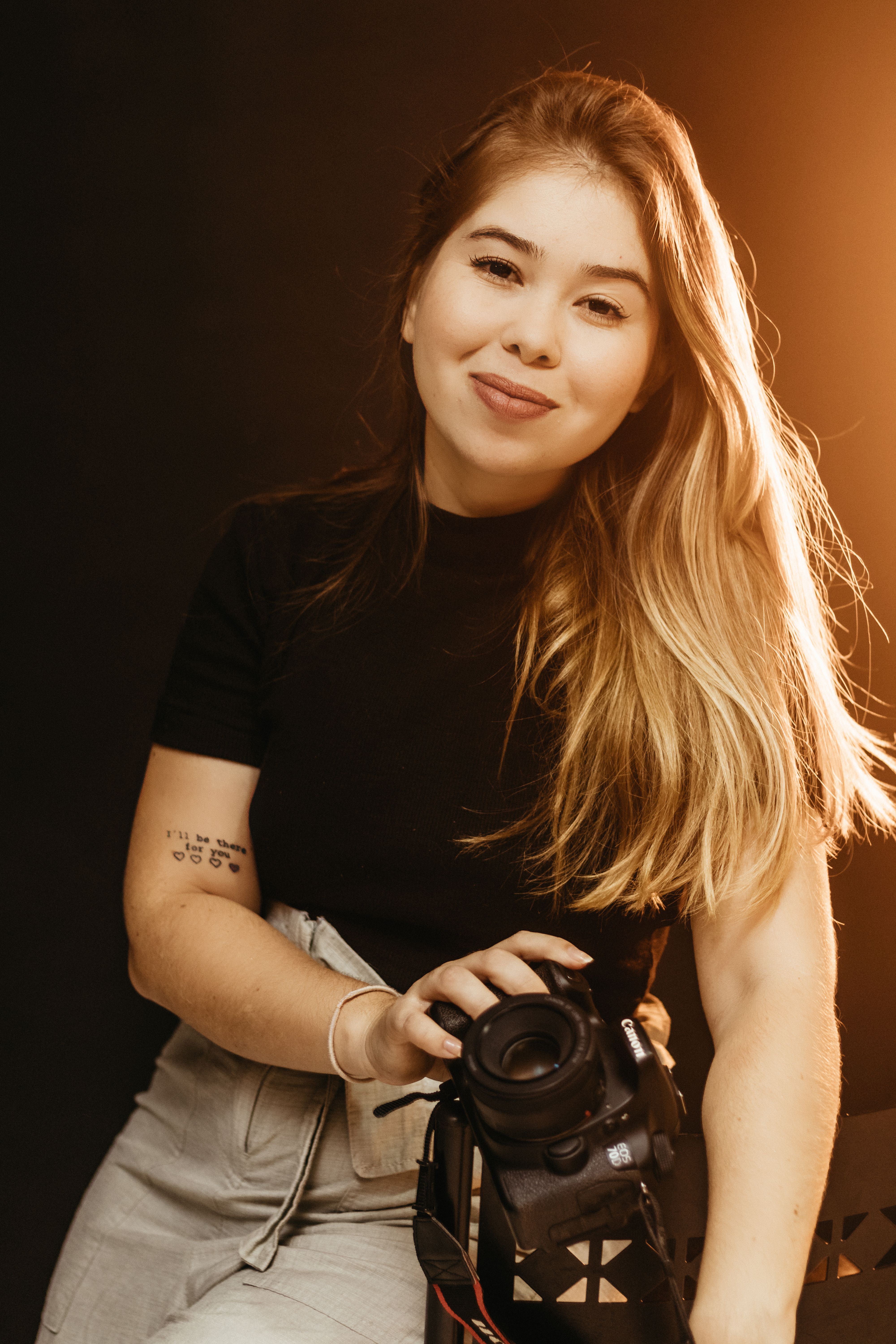 Portrait of a female photographer holding a camera in a warmly lit studio setting.