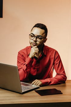 An adult male in a red shirt thinking at a desk with a laptop and tablet in a modern office setting.