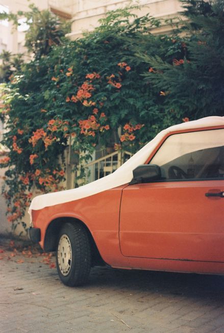 A vintage red car is covered and parked beside a wall adorned with vibrant flowers in a Bursa street.