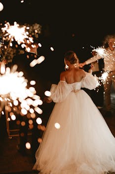 Elegant bride in off-shoulder gown enjoying a sparkler-lit wedding celebration at night.