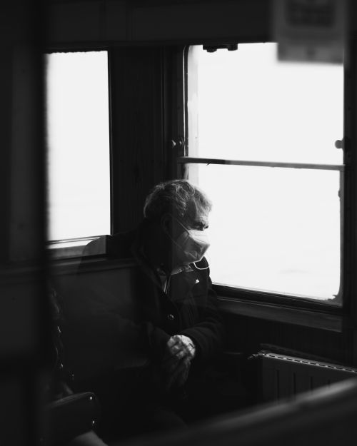 Free A black and white photo of an elderly man wearing a mask, looking out of a train window, evoking feelings of travel and contemplation. Stock Photo