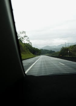 Capture of an empty road through lush greenery on an overcast day, viewed from inside a vehicle.