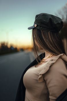 Portrait of a fashionable woman standing on a street during sunset, wearing a brown blouse and decorative hat.