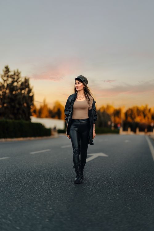 Free Stylish woman in leather outfit and flat cap walking down a road at sunset, showcasing modern fashion. Stock Photo