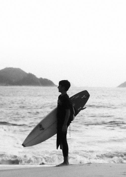 Black and White Photo of a Young Man Standing with a Surfboard on the Beach 