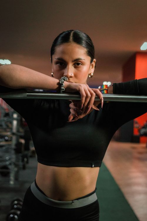 Free Portrait of a confident female athlete in gymwear leaning on a barbell in a gym setting. Stock Photo