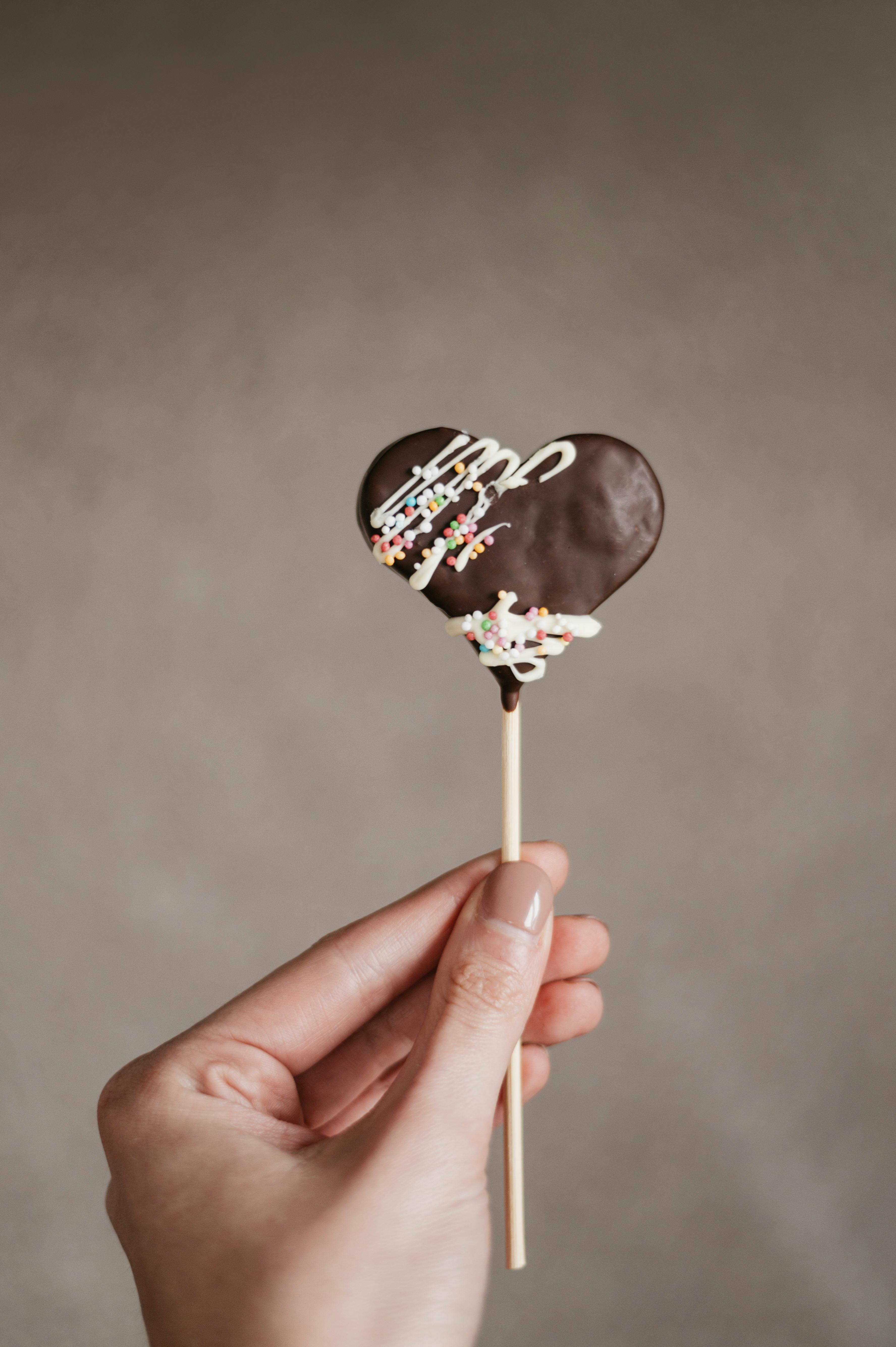 A hand holds a heart-shaped chocolate lollipop on a stick, adorned with colorful sprinkles.