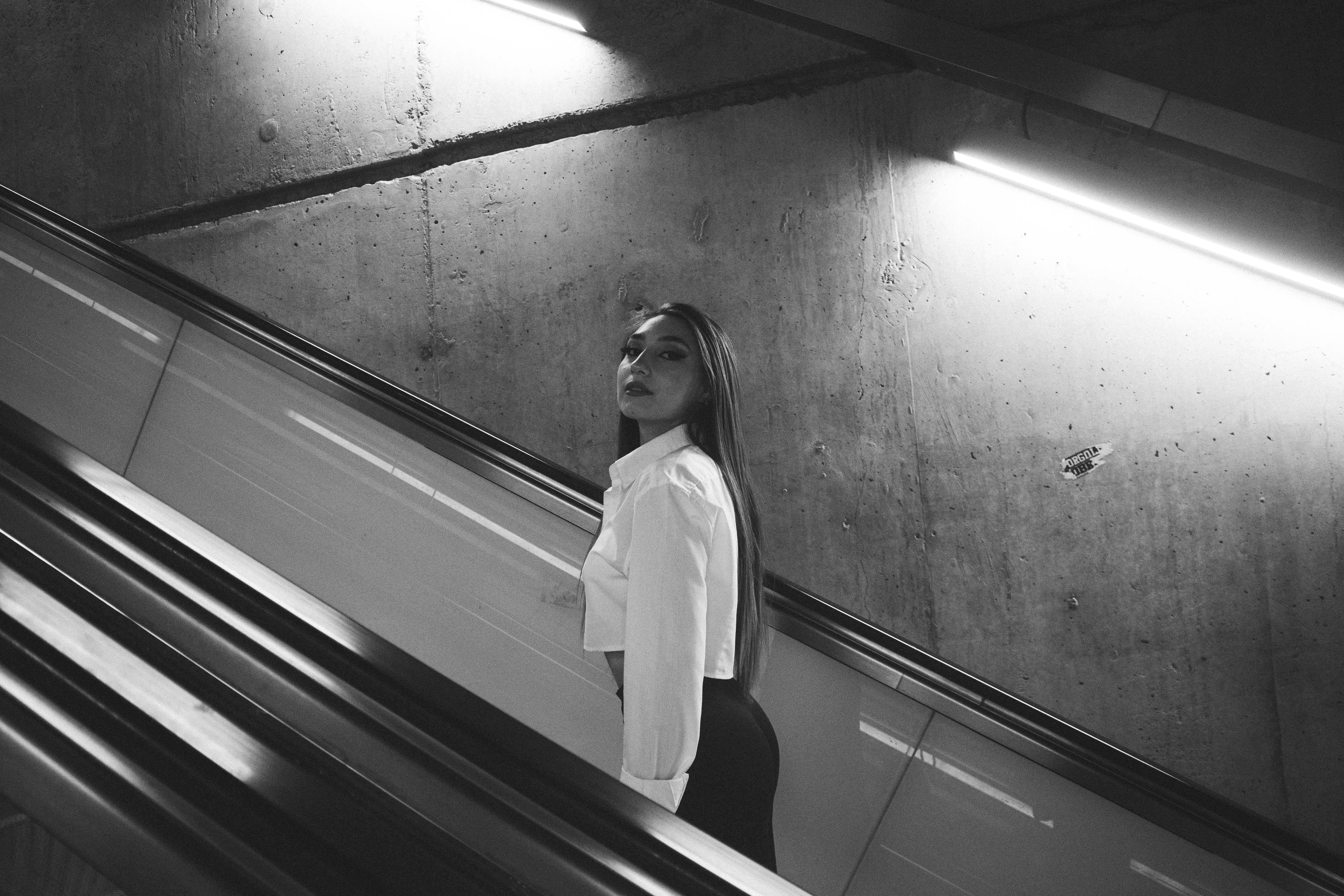 Free Black and white photo of a woman with long hair posing on an escalator indoors. Stock Photo
