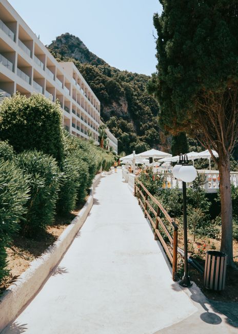Pathway leading to a Greek resort surrounded by lush greenery and mountains.