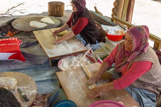 Women in headscarves making traditional Turkish flatbread outdoors on wooden boards.