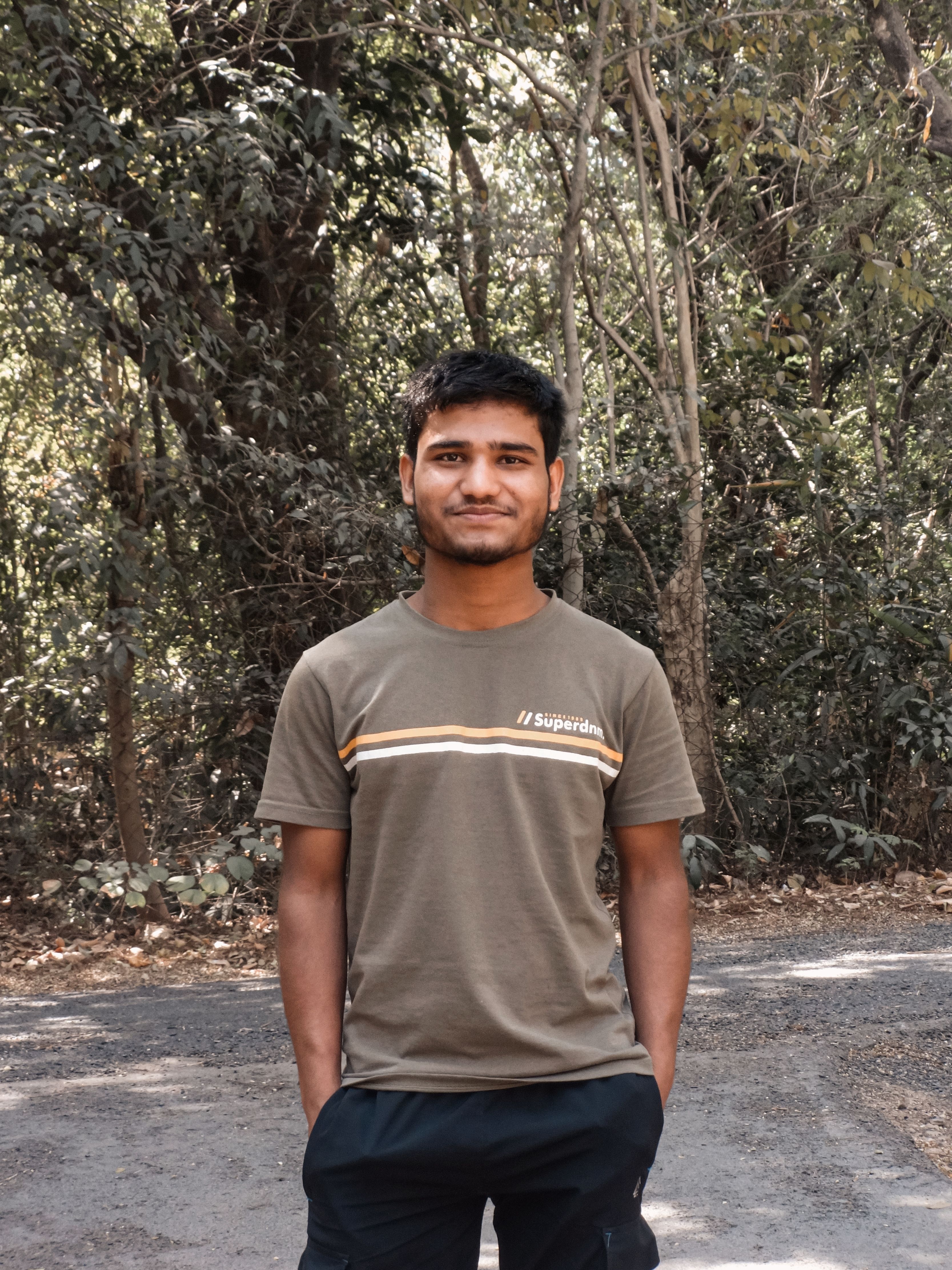 A young man standing confidently on a forest road in casual attire during daylight.