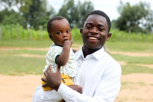 Free A joyful young man holds his baby outdoors in a park, both smiling at the camera. Stock Photo
