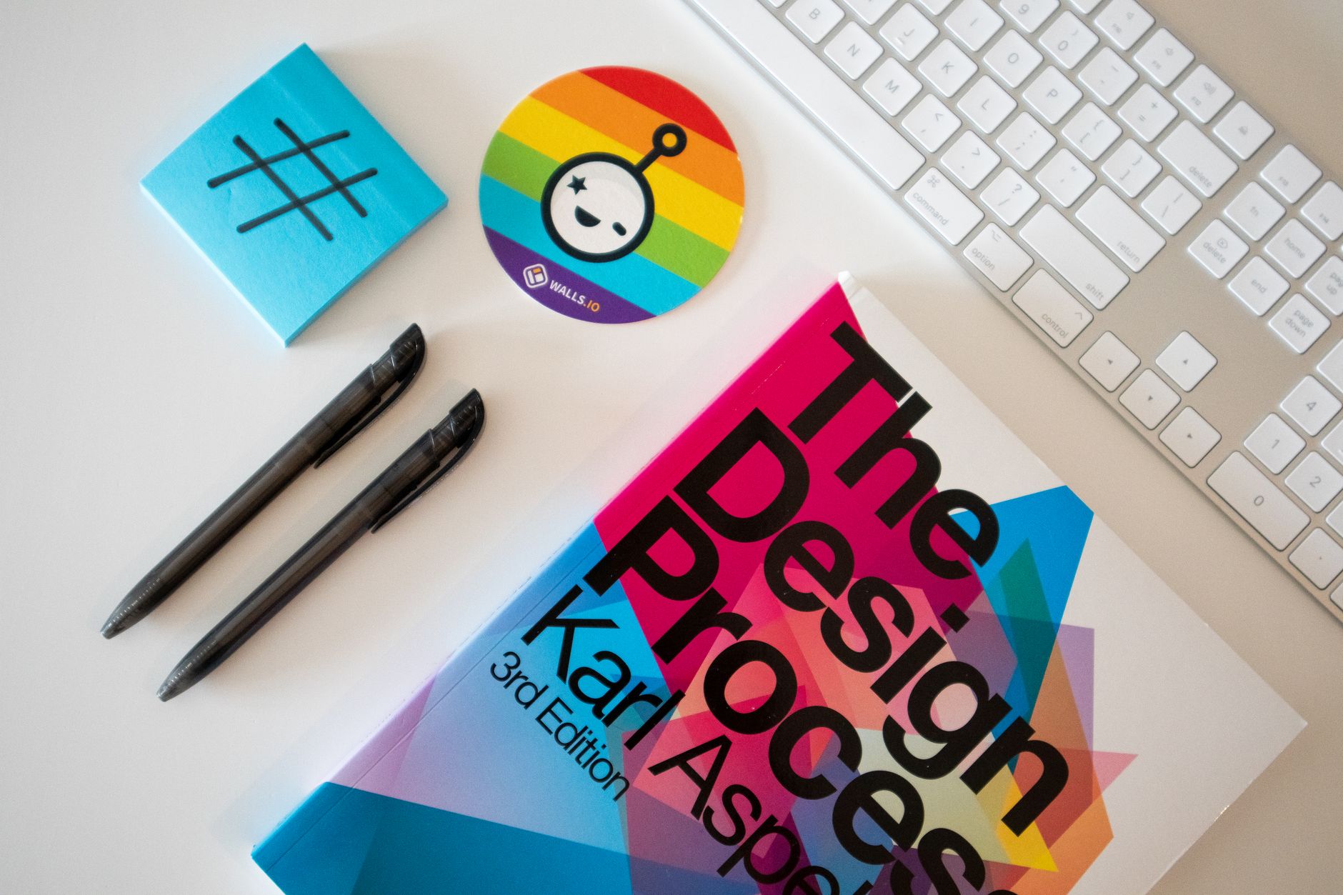 Flat lay of a creative desk setup featuring a design book, pens, and rainbow-themed sticker.