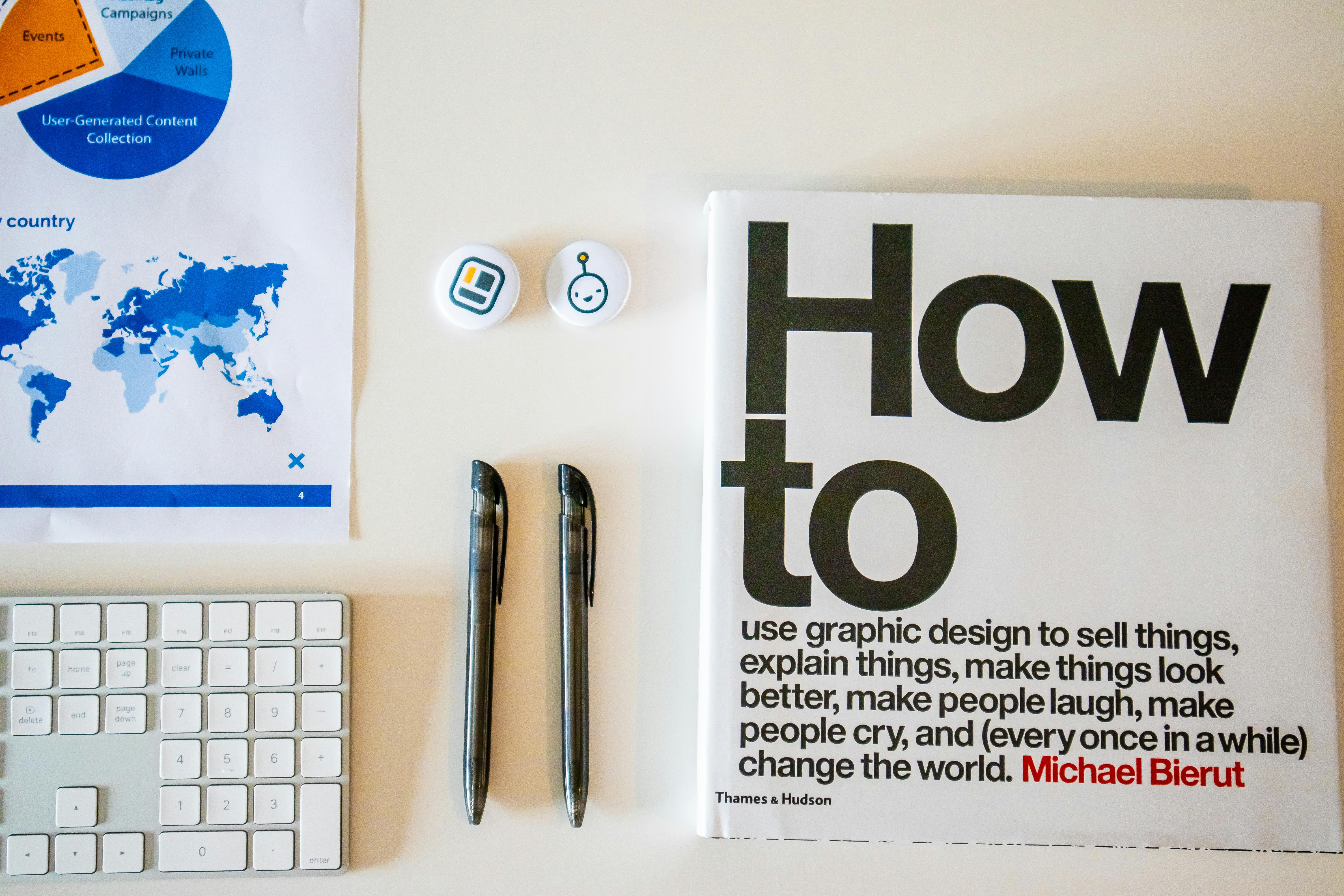 Free Flat lay of a creative workspace with 'How To' book, chart, pens, and keyboard in minimalist style. Stock Photo