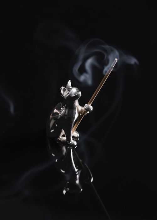 Free A close-up of a silver cat-shaped incense holder with burning incense and rising smoke against a black background. Stock Photo