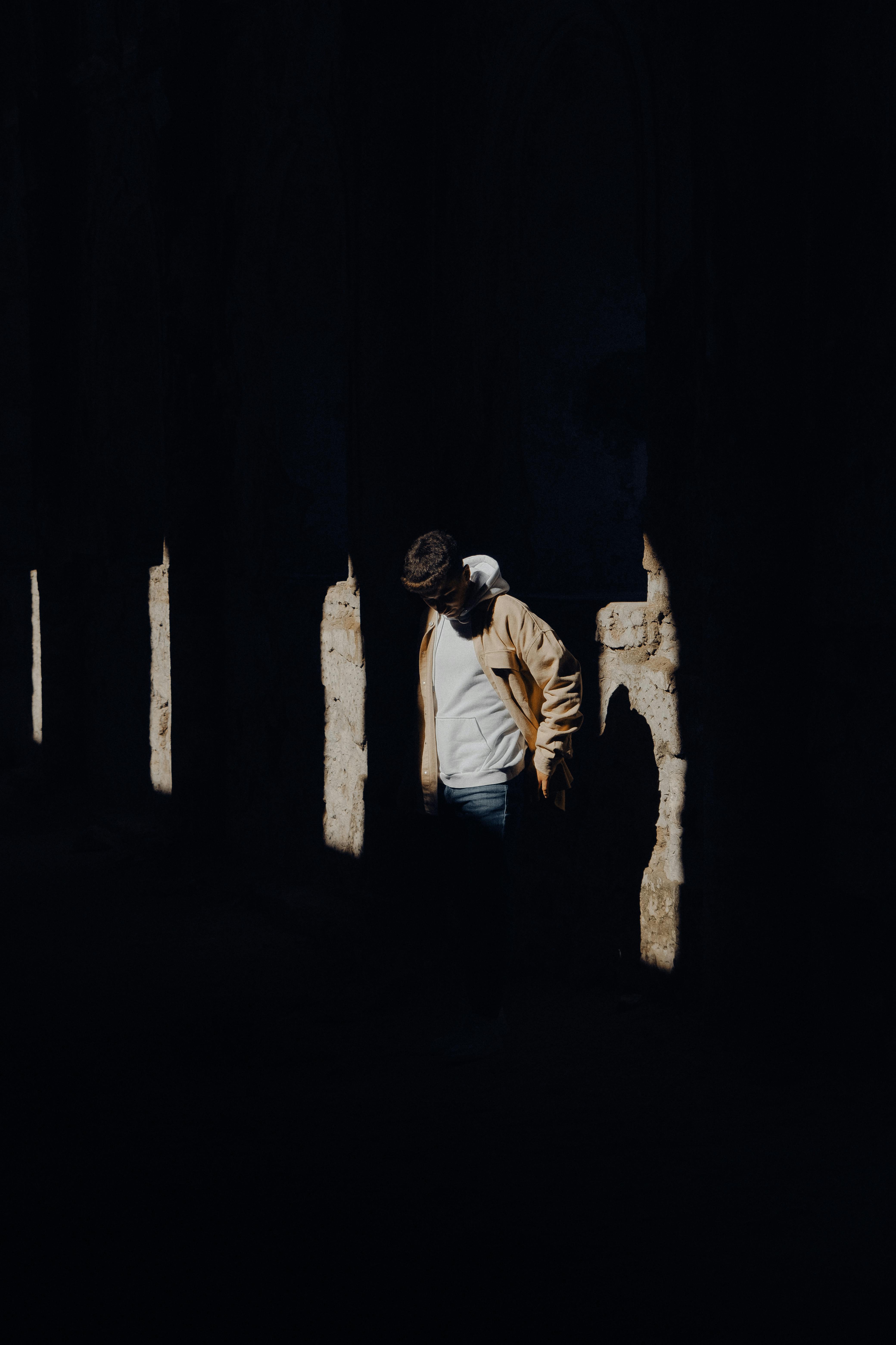 Free A lone man stands in a dark, dramatic space, partially lit by sunlight casting shadows. Stock Photo